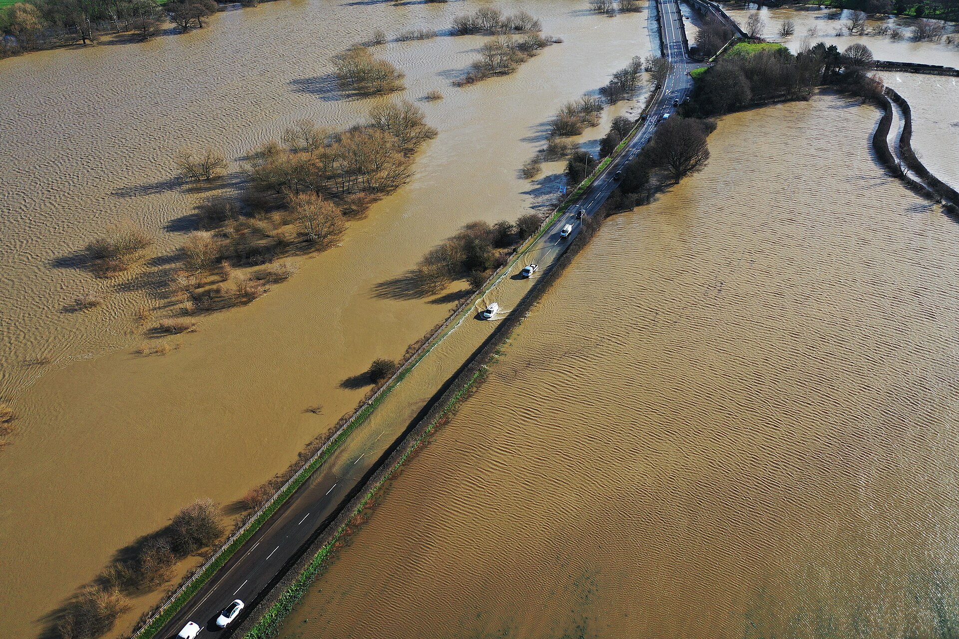 Cars driving through deep flood water on a rural road after heavy rain.