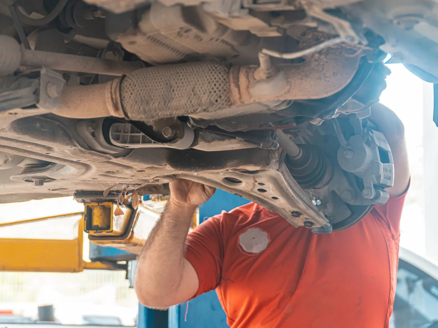 Mechanic in an orange shirt checking a car's suspension from underneath on a lift.