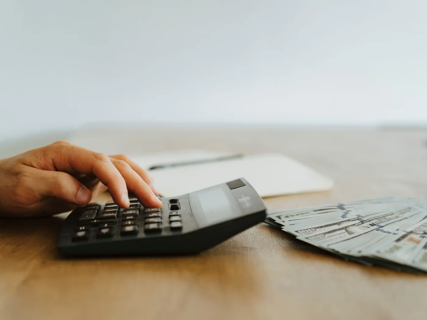 Hand using a desk calculator next to a stack of cash on a wooden table.